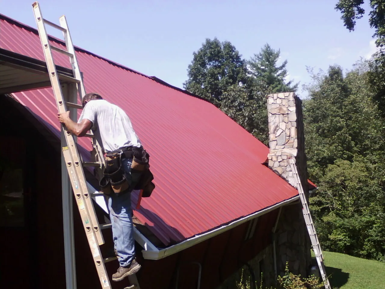 Roofer on a ladder beside a red metal roof and stone chimney — Soffit & Fascia Repair by Roofing Brothers in Norwood Young America